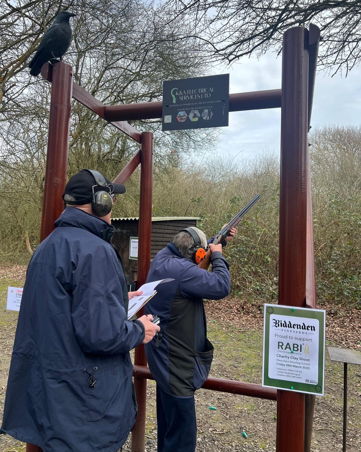 An attendee aiming at a clay pigeon, with a man standing behind him, scoring the shot.