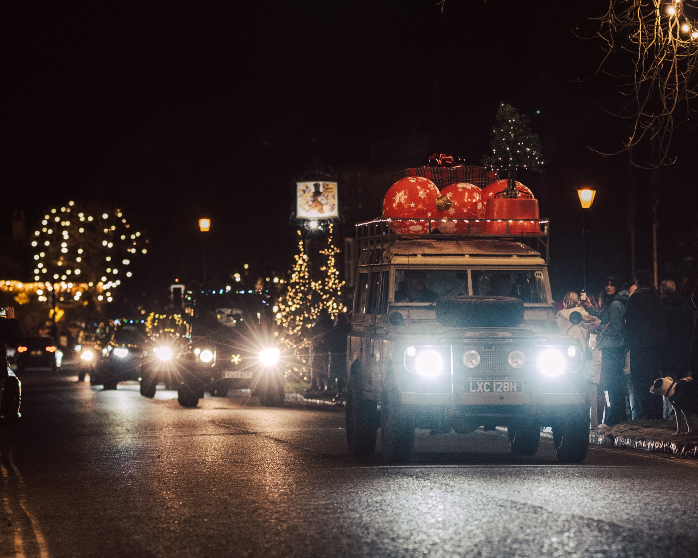 Brightly decorated Land Rover Defenders line the road as onlookers watch the festive convoy.