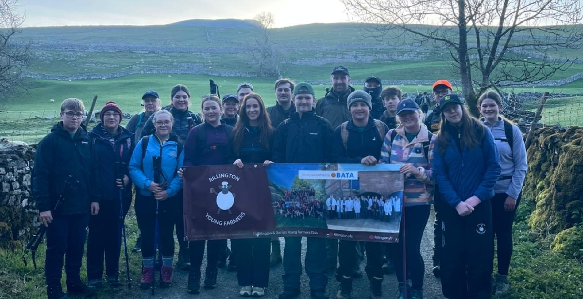 A large group from Rillington Young Farmers Club stand together on a countryside path, holding a club banner, with rolling green hills and dry stone walls behind them.