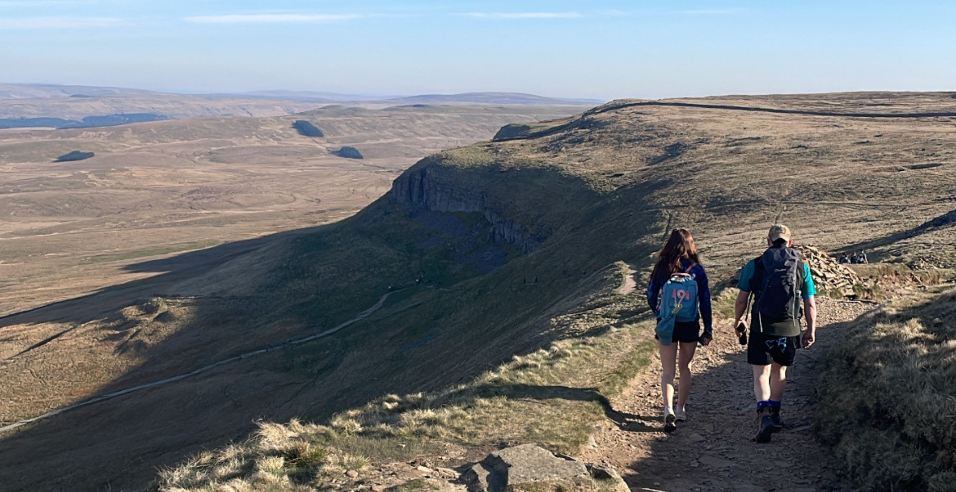 Two people walk along a high ridge path overlooking wide open moorland and cliffs, carrying backpacks under a clear blue sky.