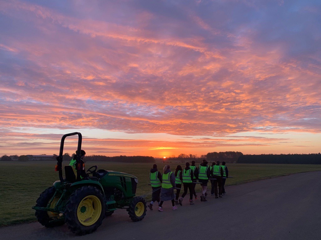 Group of people in high-visibility vests walking along a rural road at sunrise beside a tractor, with a vivid pink and orange sky overhead.