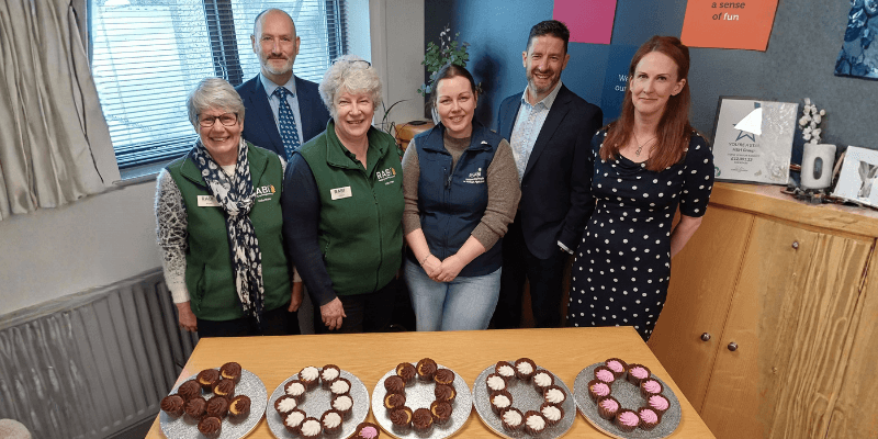 H&H-fundraiser-news Group of volunteers and supporters standing behind a table of homemade cakes arranged in patterns, smiling at the camera in a community room.