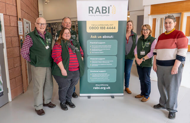 L1001008-Enhanced-NR-Edit copy (1) Group of RABI volunteers standing beside a RABI information banner displaying contact details and support services. The banner highlights practical, financial and emotional support, including the 24/7 helpline number.