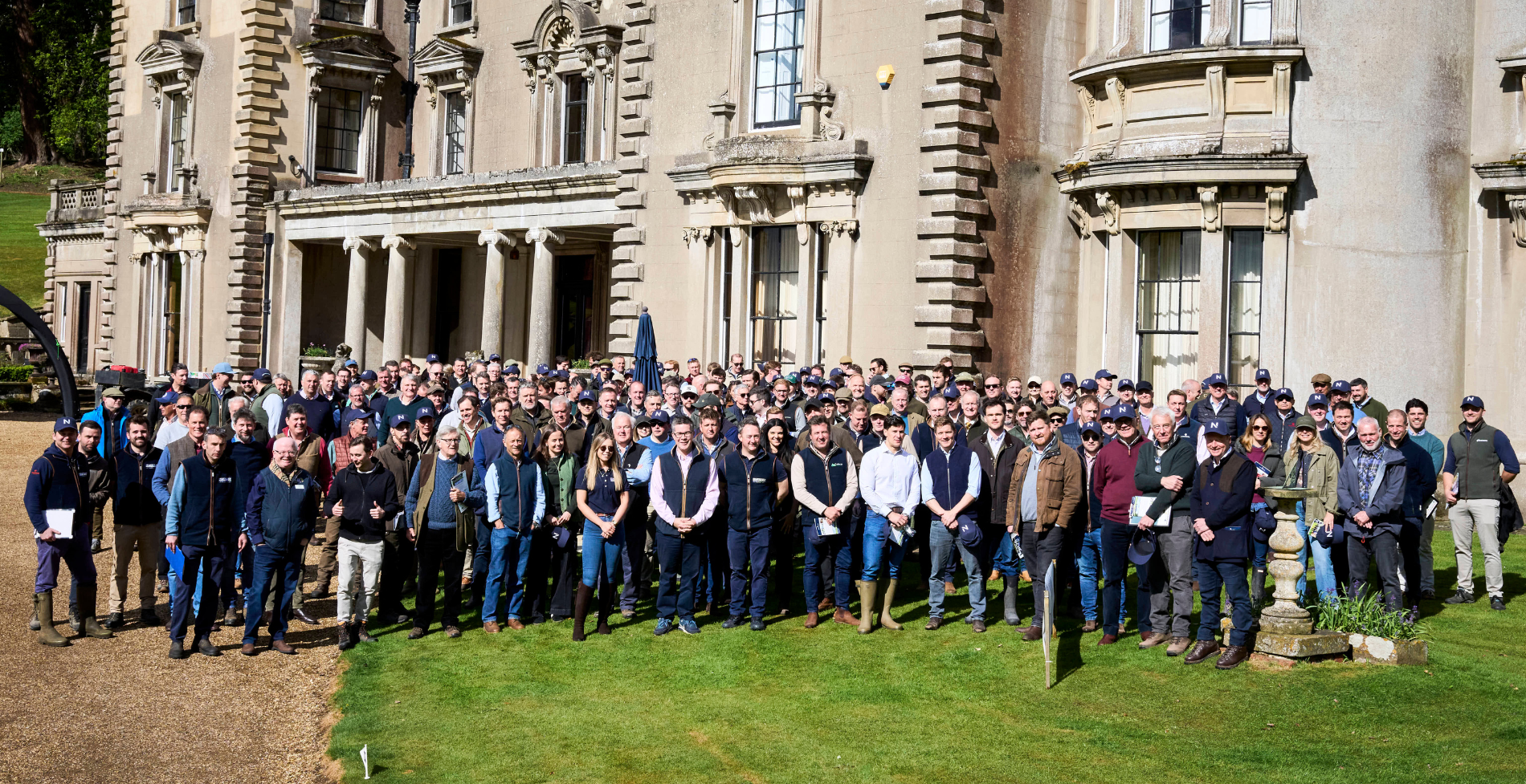 Large group of people gathered on the lawn outside a grand country house during the Newton Shoot event on a sunny day.