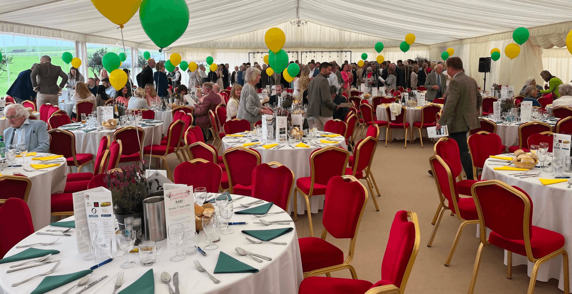 A large fundraising luncheon taking place inside a marquee. Round tables are set with white tablecloths, table settings and floral centrepieces, while guests mingle and take their seats. Red chairs and green and yellow balloons decorate the venue, creating a welcoming atmosphere for attendees supporting RABI’s County Durham Summer Luncheon fundraiser.