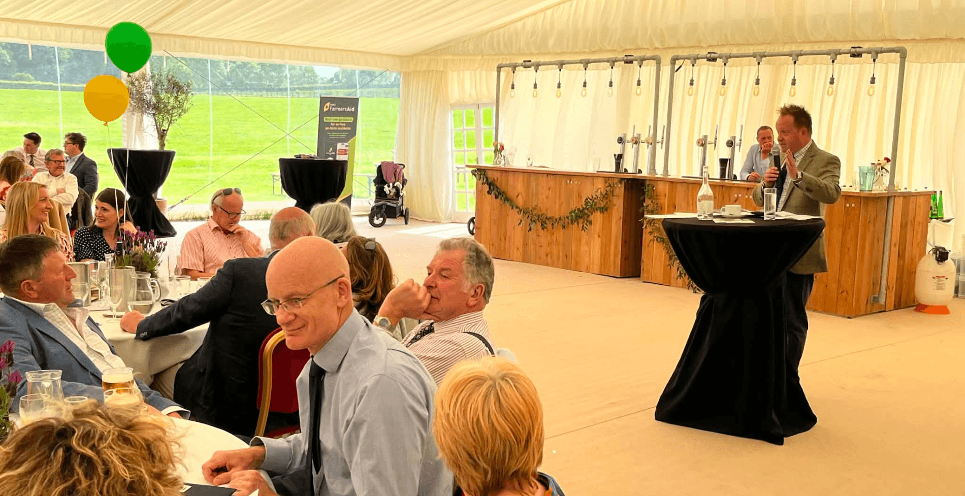 Guests attend RABI’s County Durham Summer Luncheon inside a large marquee. People are seated at round tables while others listen to a speaker addressing the audience from the front of the room. Green and yellow balloons decorate the venue, and a FarmersAid display can be seen in the background. The event brings together supporters, sponsors and members of the farming community in support of RABI’s work.