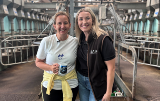 Marie and Laura Ractiffe smiling together in a dairy parlour, with Laura wearing RABI branded clothing during a farming challenge activity.