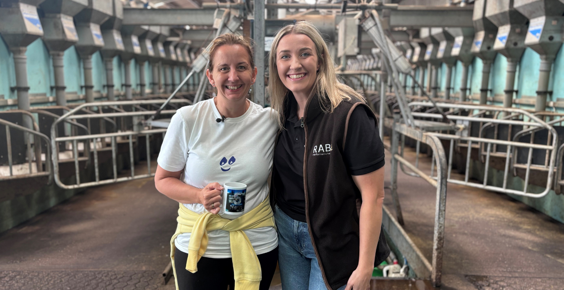 Marie and Laura Ractiffe smiling together in a dairy parlour, with Laura wearing RABI branded clothing during a farming challenge activity.