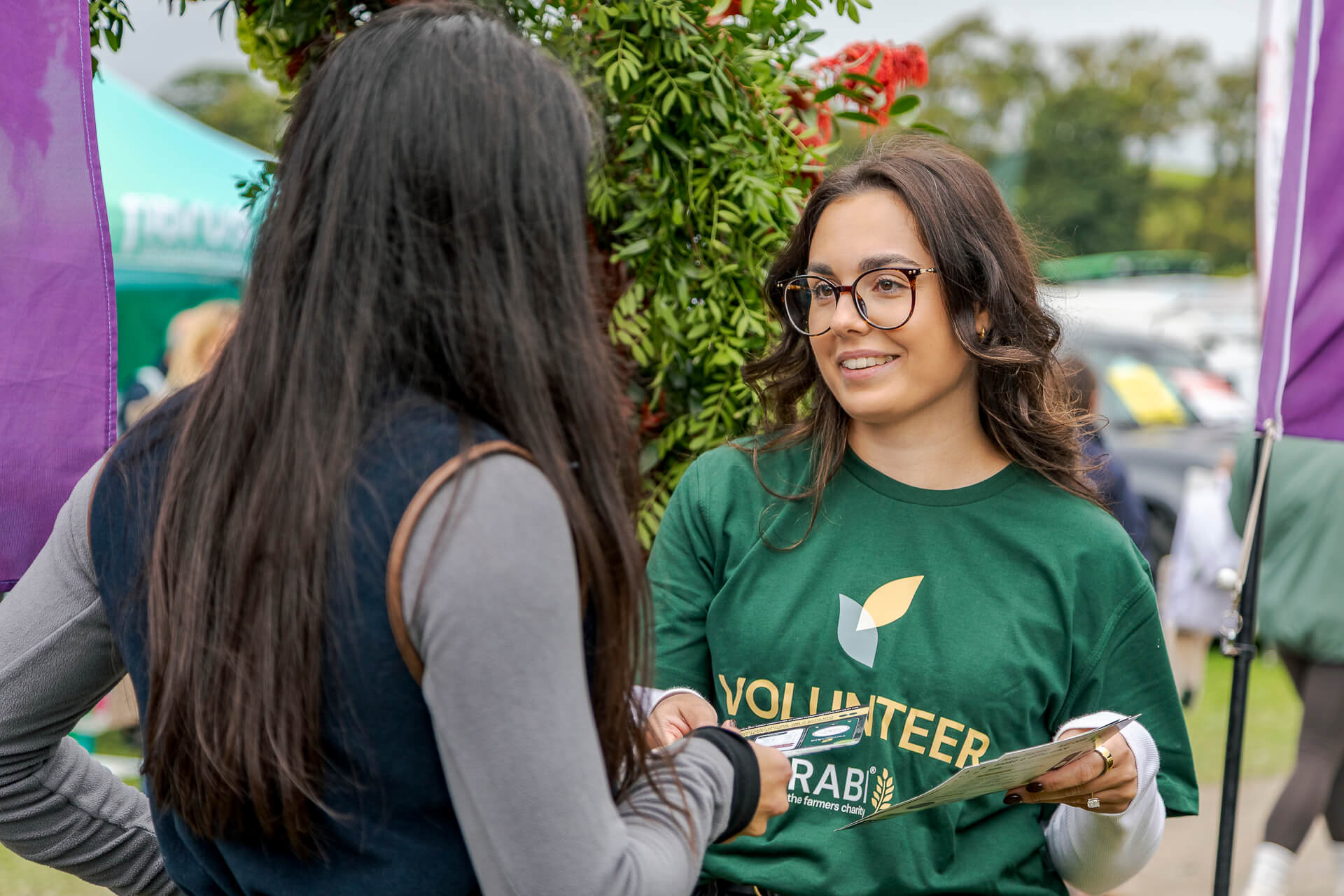 A RABI volunteer wearing a green branded T-shirt chats with a visitor at an outdoor event, holding information leaflets.