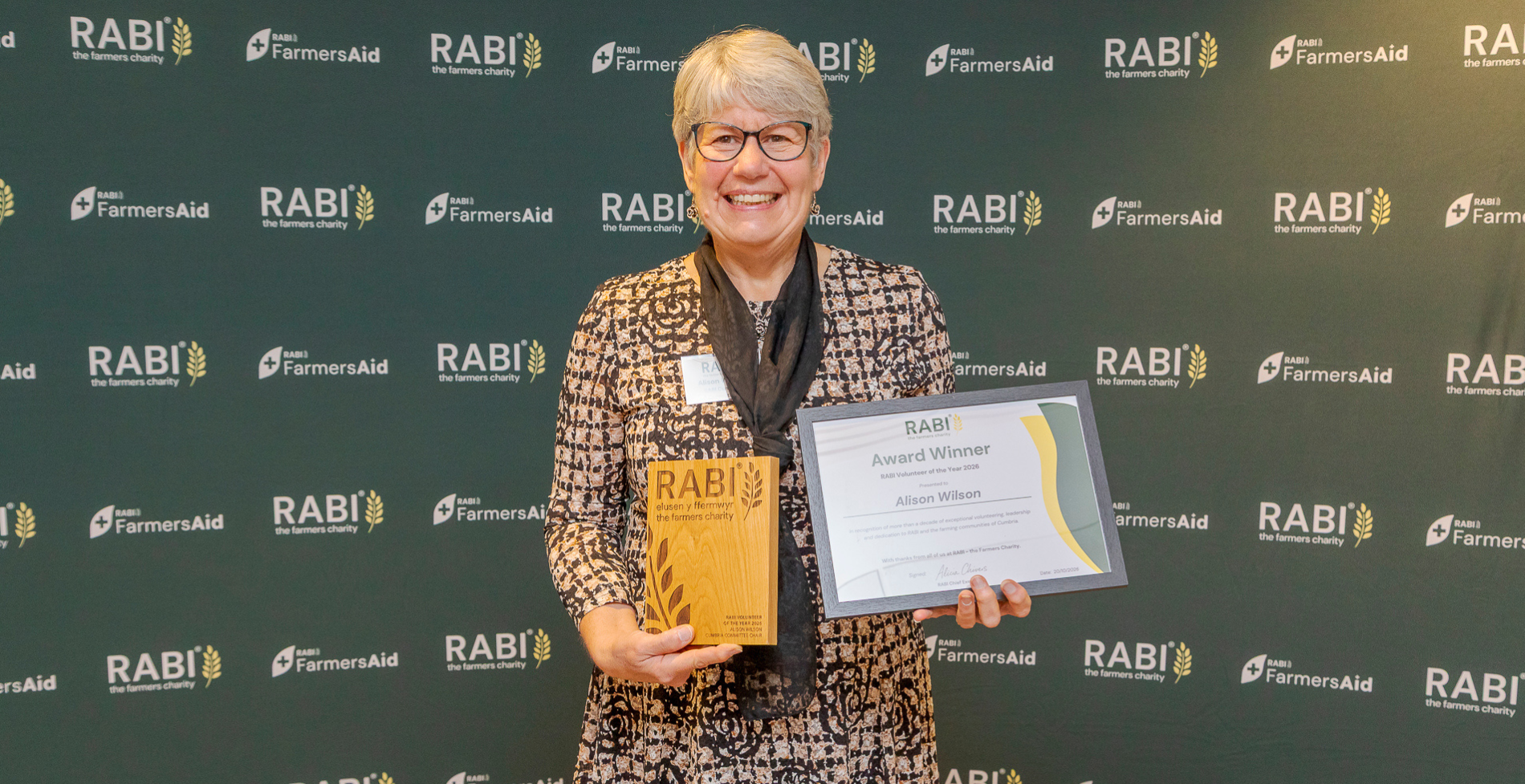 Alison Wilson smiling while holding her RABI Volunteer of the Year 2026 trophy and certificate, in front of RABI and FarmersAid branding.